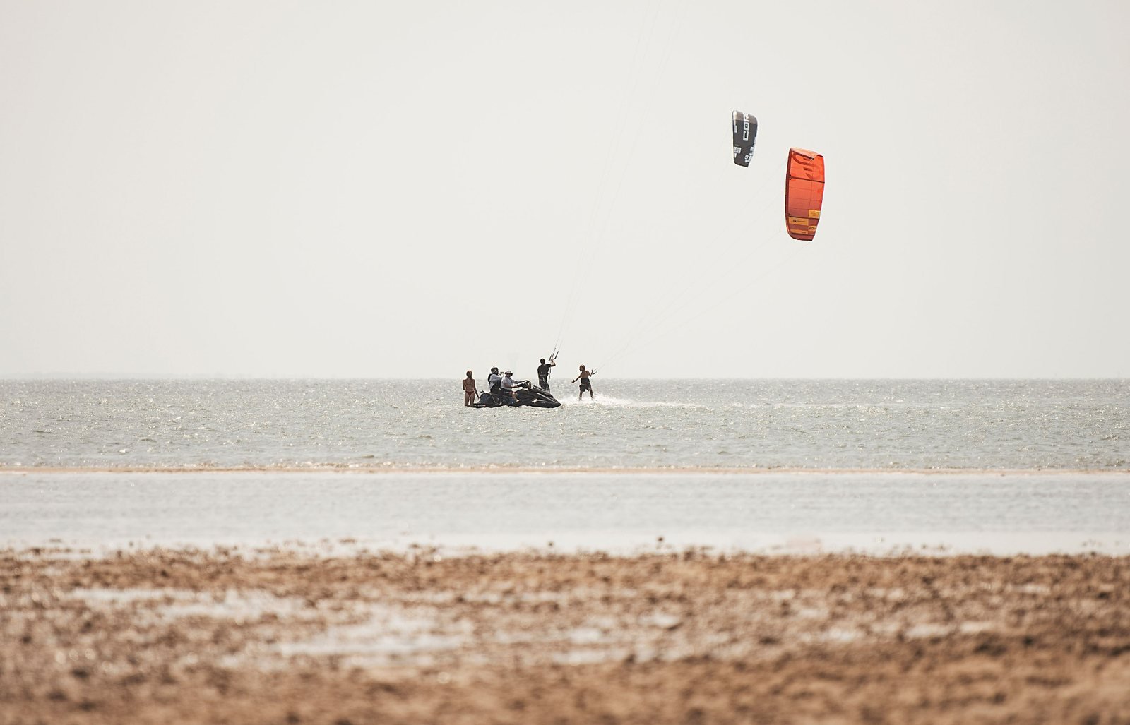 Kiteboarders gathering in shallow water beneath orange kites